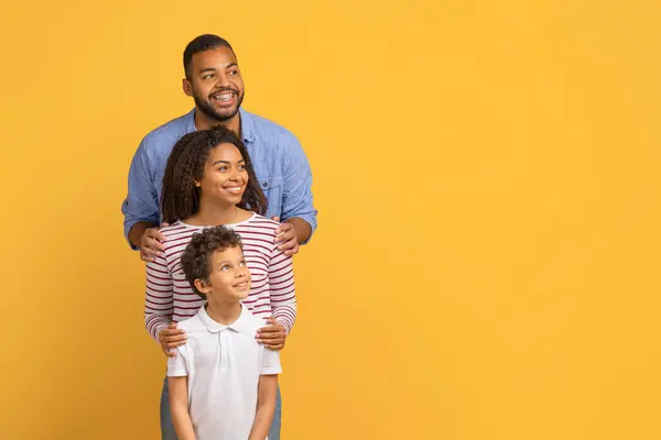 コピースペース 陽気な両親 黄色いスタジオの背景にポジショニング 新しいオファーやプロモーションを楽しんで パノラマに立っている3人の幸せな若い黒人家族 — ストック写真