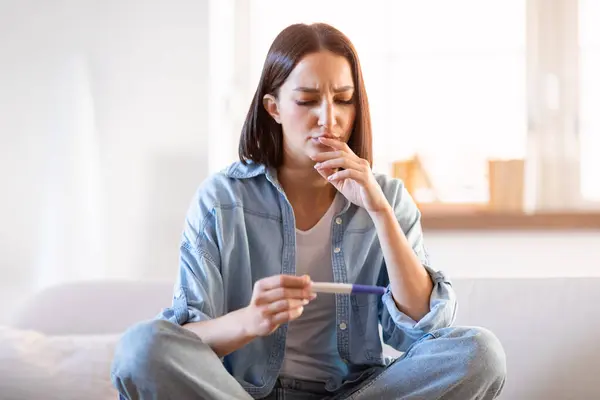 Unhappy young woman with pregnancy test sitting on couch at home, sad about negative result and infertility or unplanned pregnancies. Problems with conceiving child, female health, unwanted baby