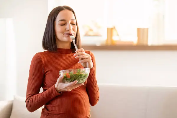 Healthy diet during pregnancy. Pregnant woman enjoying vegetable salad meal, posing with bowl and fork at home, sitting on couch indoor. Expecting and eating healthy food concept