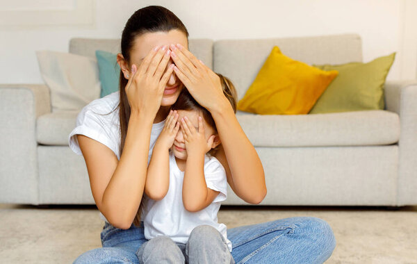 Мама и ребенок играют в пикабу дома, молодая мать и маленькая дочь сидят на полу, закрывая глаза и улыбаясь, веселятся вместе в хорошо оформленном интерьере гостиной,