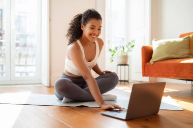 Sportif bayan, bilgisayarını, modern oturma odasındaki spor paspasının üzerinde oturarak online yoga egzersizi için hazırlıyor. Sağlık ve günlük spor motivasyonu kavramı