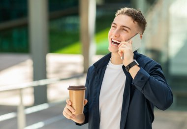 Deniz mavisi ceketli neşeli genç bir adam akıllı bir telefonla konuşuyor, elinde tek kullanımlık bir kahve fincanı var, arka planda modern bir binayla açık bir şehir ortamında duruyor.