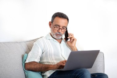 Son sınıf öğrencisi cep telefonuyla konuşuyor evdeki oturma odasında dizüstü bilgisayarda çalışıyor, bilgisayardaki interneti incelerken iletişim kuruyor, evdeki koltukta otururken gözlük takıyor.