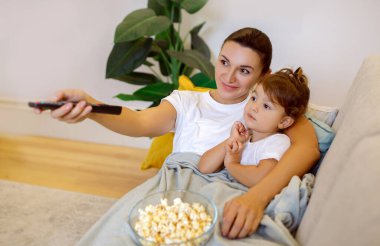 Aile eğlencesi. Küçük kızıyla TV 'de film izleyen gülümseyen genç bir anne, çocuğunu kucaklayan bir anne, patlamış mısırla koltukta oturuyor ve uzaktan kumandayla uzaktan kumandayı tutuyor, yakın çekim.