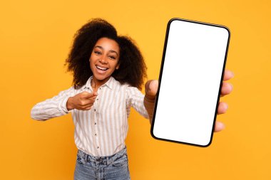 Kıvırcık saçlı neşeli genç kadın boş akıllı telefon ekranını hevesle işaret ediyor. Uygulama promosyonu için ideal, parlak sarı arka plan, model.