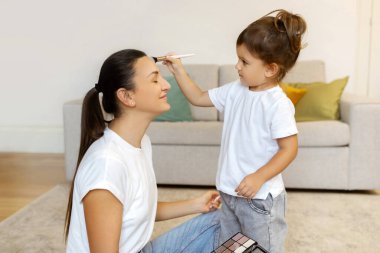 Sevimli küçük kız, anne evde oynarken makyaj yapıyor, sevimli anaokulu kızı ve genç annesi birlikte eğleniyor, kıkırdayarak eğleniyor ve eğleniyor.