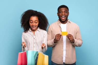 Çoklu alışveriş çantaları ve altın kredi kartıyla neşeli Afro-Amerikan çifti, eğlenceli perakende terapi gününü kutluyorlar, canlı mavi arka planda, mutluluk ve tüketici sevinci yayıyorlar.