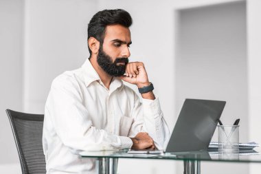 Düşünceli Hintli profesyonel, sakallı, parlak cam bir masadaki bilgisayarının üzerinde kafa yoran, çağdaş bir ofiste odaklanmış problem çözmeyi somutlaştıran