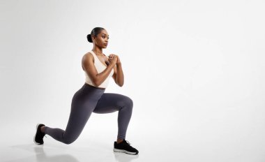 Afro-Amerikalı fitness kadın bacaklarını açıyor beyaz stüdyo arka planında ileri doğru hamleler yapıyor, egzersiz rutininden zevk alıyor, yan bakıyor. Tam boy çekim, boş alan