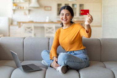 Kredi kartı tutan ve kameraya gülümseyen mutlu Hintli kadın internetten alışveriş yapmaya hazırlanan neşeli ester kadın evdeki rahat oturma odasındaki kanepede dizüstü bilgisayarıyla oturuyor.