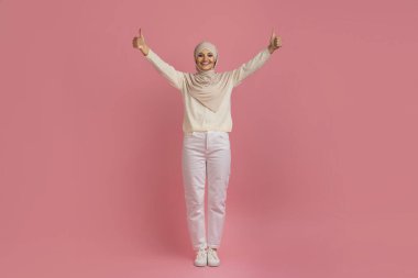 Mutlu Müslüman kadın tesettürde el kaldırıyor ve kameraya baş parmağını kaldırıyor, başörtüsü takmış gülümseyen Arap kadın onaylama işareti yapıyor, pembe stüdyo arka planının başında duruyor, tam boy, kopyalama alanı.