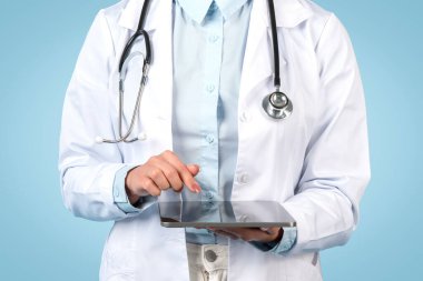 Laboratuvar önlüklü kadın doktorun tıbbi kayıtları güncellemek ya da kontrol etmek için dijital tablet kullanırken çekilmiş resmi, boynunda stetoskop, mavi arka plan.