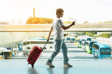 Yandan bakan genç Asyalı turist elinde akıllı telefonuyla havaalanının önünden geçiyor, bavulunu taşıyor, biniş bilgilerini internetten kontrol ediyor, güneş parlaması, kopyalama alanı.