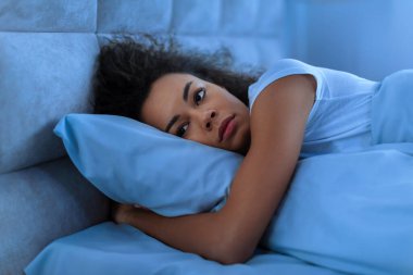Depresyondaki Afro-Amerikan kadın evde yatağında tek başına uzanıyor, modern yatak odasında yalnızlık ve uykusuzluk çekiyor. Uykusuzluk konsepti. Düşük ışık