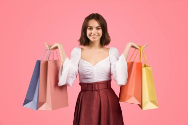 Beyaz bluz ve kestane rengi etek giyen gülümseyen genç Avrupalı kadın, mutluluk ve güven saçıyor, canlı pembe bir arka planda renkli alışveriş torbaları tutuyor, stüdyo