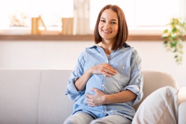Avrupalı hamile bir kadın mutluluk saçıyor, rahat bir şekilde kanepeye oturuyor, karnına hafifçe dokunuyor, açık oturma odasında bebeğini bekliyor, kameraya gülümsüyor. Boş boşluk
