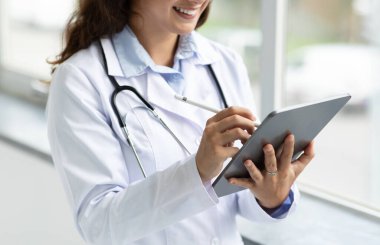 Doktor elleri dijital tablet kullanıyor, kadın doktorunun hastayla online randevusu var, klinik hastanesinin penceresinin yanında duruyor. Modern sağlık teknisyenleri