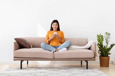 Milenyum Asyalı gülümseyen bayan akıllı telefon tutuyor, rahat oturma odasında, ev içinde kanepede oturuyor. Filipinli kadın çevrimiçi kitap veya blog okuyor cep telefonu, kopyalama alanı, tam uzunluk
