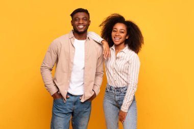 Neşeli genç siyah adam ve kadın, kadın kolları erkeğin omzunda, güneşli sarı bir arka planda kameraya gülümsüyorlar.