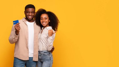 Gülümseyen genç Afrikalı Amerikalı çift, mutlu sarı arka plana karşı güvenle kredi kartı gösteriyor ve serbest alanı işaret ediyor, finansal özgürlüğü veya satın alma onayını simgeliyor.