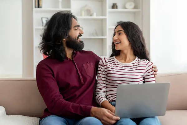 Excited millennial indian man and woman sitting on sofa in cozy living room interior, looking at each other and smiling, using laptop at home, shopping online, choosing furniture