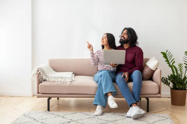 ポジティブな陽気なミレニアルインド愛好家はソファに座って 自宅でラップトップを使用して 白い壁の背景の上に空白のコピースペースを指し 素敵なウェブサイトを示し オンラインオファー — ストック写真
