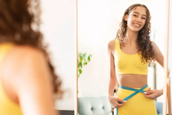 Smiling caucasian teen girl in sportswear measuring waist with tape, checking inches lost around her belly, feeling proud and happy with her weight loss results, looking at mirror at home