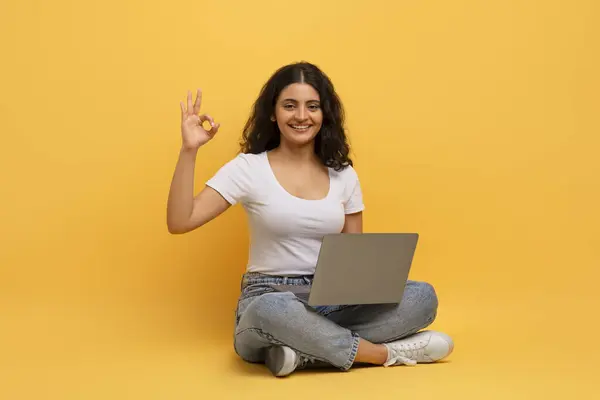 ポジティブな陽気な好奇心の若いインドの女性学生は 彼女の膝の上にラップトップコンピュータと床に座って 大丈夫なジェスチャーを示し オンラインで勉強し コースを推奨 黄色の背景 — ストック写真