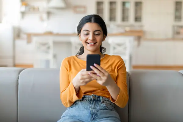 Beautiful young indian woman using her smartphone while sitting on couch, happy eastern female comfortably lounging on gray sofa in bright, contemporary living room setting, browsing mobile app