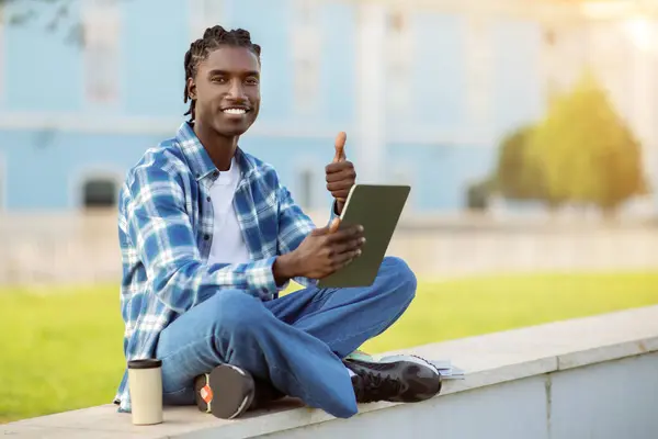 若い黒人学生の男は デジタルタブレット上の教育ウェブサイトを閲覧し 電子学習コンテンツを承認するために親指を与える 勉強することを躊躇 大学のキャンパス外に座って — ストック写真