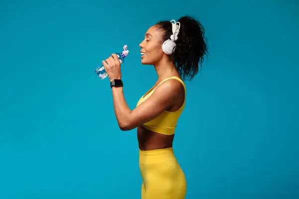 stock image African American fitness lady with earphones sipping from a water bottle, in yellow wear, on blue studio backdrop, symbolizing active lifestyle and healthy hydration during workout. Side view