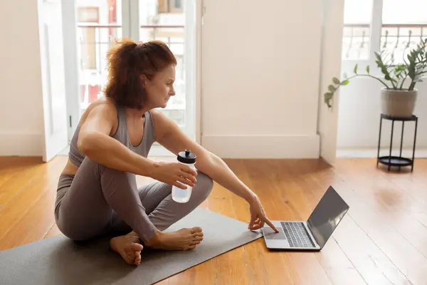 Online Fitness Tutorials. Beautiful Senior Lady Preparing For Training At Home, Opening Laptop Or Browsing Videos With Exercises, Elderly Woman Sitting On Yoga Mat In Living Room, Free Space