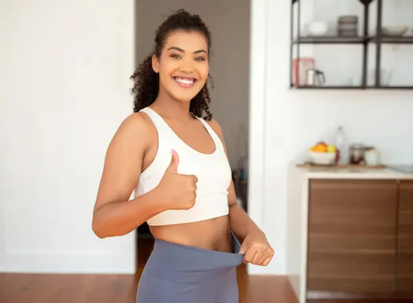 Successful weight loss. Athletic young woman in fitwear gesturing thumbs up while pulling waist of fitness pants, smiling to camera happy about slimming progress at home interior