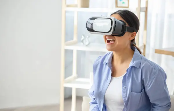 Young asian woman in VR headset touching air during virtual reality experience at home. Pretty chinese lady exploring artificial computer world or playing video game indoors, copy space