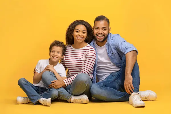 床に座って笑顔でカメラに向かって座っている 幸せな若いアフリカ系アメリカ人の両親と彼らの男性の子供が抱擁し 結合し 黄色の背景に隔離されたポーズ — ストック写真