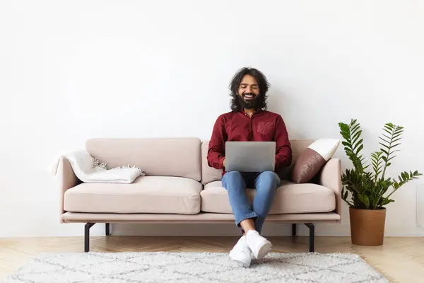 Handsome positive millennial indian man using laptop at home, sitting on couch in cozy living room interior over blank white wall background with blank copy space. Freelance, remote job