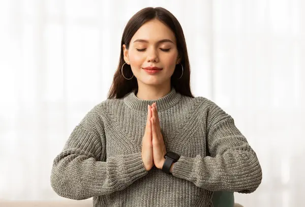 Stress-relief, mindfulness, meditation. Portrait of peaceful young woman meditating with closed eyes, sitting on couch next to window, holding hands in namaste gesture, closeup