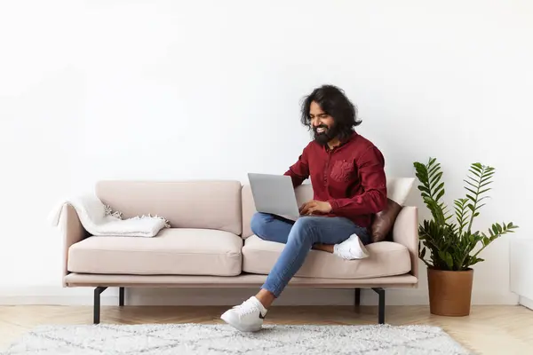 Cool bearded long-haired millennial eastern guy hipster sitting on couch at home, using computer laptop. Indian man working remotely, chatting with friends, websurfing, copy space