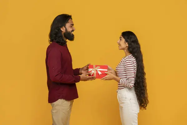 かわいい陽気な若い東部のカップルは 黄色のスタジオの背景に贈り物を交換しています 横のビュー 赤いギフト用の箱を保持し お互いに微笑んでいるインドの男と女性 聖バレンタインデーのお祝い — ストック写真