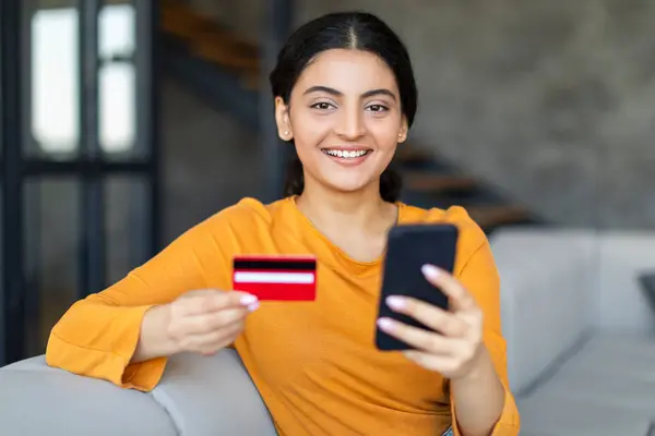オンライン決済を手元で行うクレジットカードを持つ陽気なインド人女性 自宅で居心地の良いリビングルームのインテリアに座っている間 彼女の携帯電話を使用して幸せな東部の女性 無料スペース — ストック写真