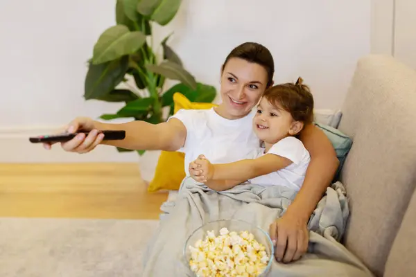Family Movie Time. Mother and little daughter with snacks relaxing on couch and watching tv, happy mom and cute preschooler female child having fun together, lying under blanket and eating popcorn