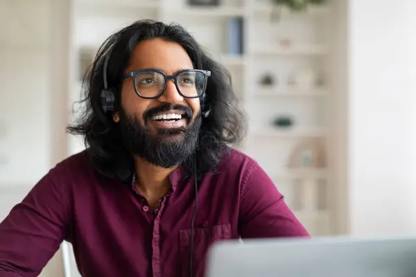 Indian freelancer guy with glasses and headset sitting at desk with laptop and looking away, dreamy happy eastern man thinking about something good while working in home office, closeup shot