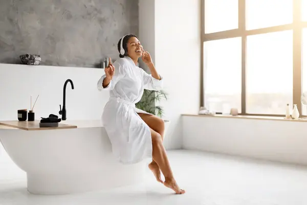 身穿白色浴衣的快乐黑人女性一边在奢华的浴室里一边听着带耳机的音乐一边跳着舞 积极的非洲裔美国女性坐在浴缸上 欣赏着最喜欢的歌曲 — 图库照片