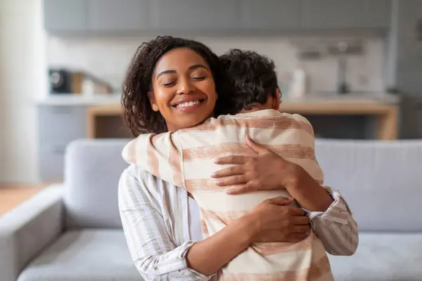 微笑着的黑人母亲在家里热情地拥抱着她十几岁的儿子 快乐的年轻的非洲裔美国女人拥抱着男孩 充满爱心的父母在舒适的客厅里表现出爱心和安慰 — 图库照片