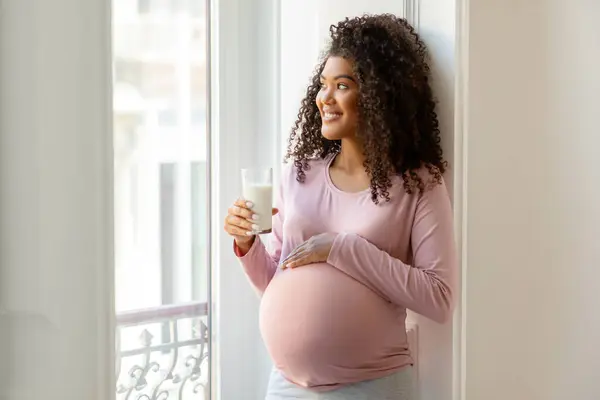 ハッピーブラック妊娠中の女性の肖像画 家で窓の近くに立って腹に触れながら 期待するアフリカ系アメリカ人女性 健康なカルシウム飲料を楽しむ コピースペース — ストック写真