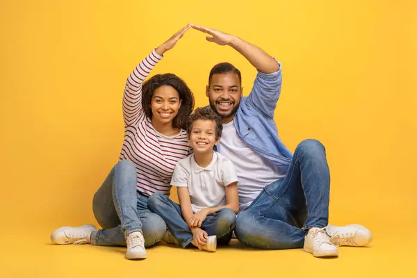 Family Protection. Loving African American Parents Making Roof Of Hands Above Their Little Son, Caring Black Mother And Father Bonding With Male Child While Sitting Together On Yellow Background