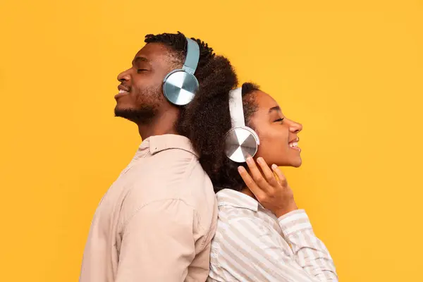 Relaxed young black couple standing back-to-back, each wearing headphones, lost in music with content smiles on a sunny yellow background, side view