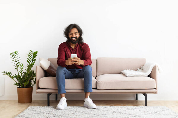 Веселый бородатый длинноволосый молодой индиец в повседневной одежде сидит на диване в уютном интерьере гостиной дома, используя телефон, переписываясь с друзьями, тестируя новое мобильное приложение, копируя пространство