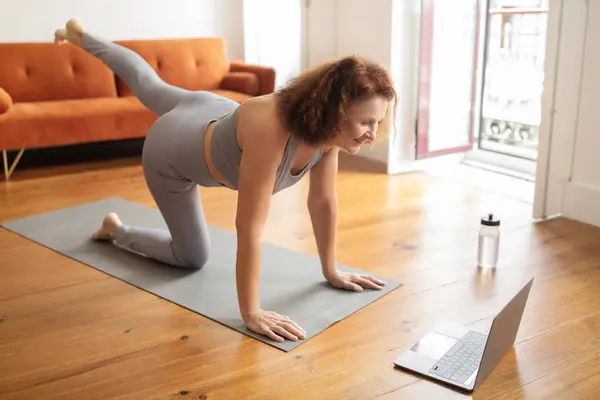 Senior woman exercises with online class at home. Active elderly lady in sportswear following fitness workout lesson, performing leg lift exercise on yoga mat, training in living room interior