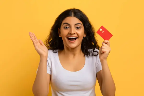 彼女の赤いプラスチックの銀行のクレジットカード ジェスチャーと笑顔 黄色の背景を示す白いTシャツを着ている感動的なかなり若いインドの女性 イージーバンキング 高速ローン — ストック写真